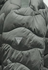 Σκούρο γκρι μπουφάν με καπιτονέ σχέδιο κυμάτων, λεία γυαλιστερή υφή και τριγωνική ετικέτα με το λογότυπο Guess στο στήθος.