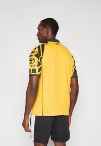 Κίτρινη αθλητική φανέλα με μαύρα μανίκια με μοτίβα, με μαύρο γιακά και λογότυπα. Φορεμένη με μαύρο σορτς, ορατές πλευρικές λευκές λεπτομέρειες.