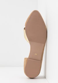 Chaussure plate en daim beige avec un bout pointu, semelle en caoutchouc avec des rainures pour l'adhérence, taille marquée 37, placée debout contre un coin blanc.