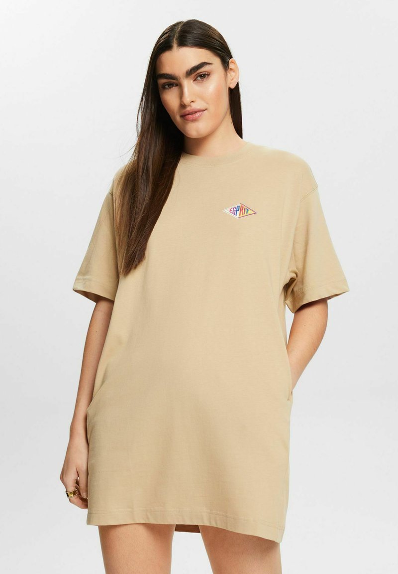 Beżowa bawełniana sukienka t-shirtowa z krótkimi rękawami, z małym logo graficznym na lewej piersi. Luźny krój z bocznymi kieszeniami.