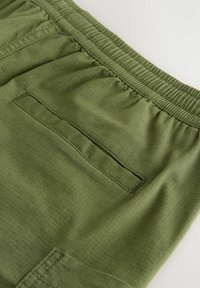 Gros plan sur un pantalon à ceinture élastique verte avec une poche arrière horizontale et un motif de tissu texturé.