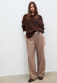 Brauner übergroßer Pullover mit cremefarbenem Ausschnitt, kombiniert mit beigen Hosen mit weitem Bein. Braune Schuhe, glattes Material, minimalistische Akzente.