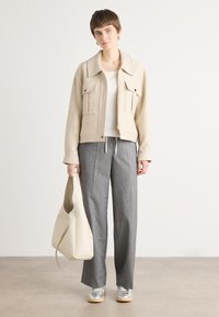 Veste beige courte avec deux poches avant, associée à un pantalon gris à rayures. Le modèle tient un sac couleur crème et porte des baskets argentées.