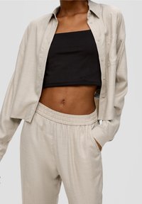 Beige button-up shirt with long sleeves, worn open over a black crop top. Features relaxed fit pants with an elastic waistband and pockets.