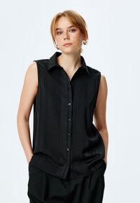 Maglietta nera senza maniche con bottoni frontali, righe verticali e colletto. Tessuto liscio, vestibilità sartoriale e una leggera lucentezza.