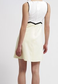 Hihaton A-linjainen mekko valkoisena ja vaaleankeltaisena, jossa on musta korostussauma ja sileä pinta, pyöreä pääntie ja takana vetoketju.