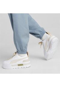 Białe trampki z beżowymi akcentami i teksturowaną, podwyższoną podeszwą. Posiadają sznurowany design oraz małe złote logo z boku.