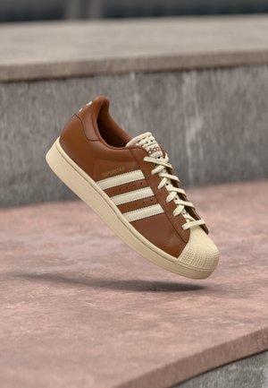 Brauner Ledersneaker mit beigen Akzenten, der eine strukturierte Zehenkappe, drei weiße Streifen an der Seite und cremefarbene Schnürsenkel aufweist.