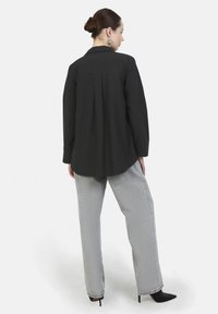Zwarte button-up shirt met een kraag en een plooitje achteraan, gecombineerd met losse grijze broek. Het model draagt puntige zwarte hakken.