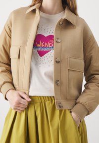 Brązowa kurtka bomberka z gładkiego materiału, z kołnierzykiem, zapinana na guziki i wyposażona w dwie przednie kieszenie. Nosi ją na białej koszulce z nadrukiem i żółtej spódnicy.