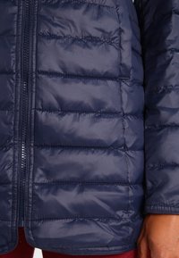 Primer plano de una chaqueta acolchada azul marino con cremallera visible y bolsillo, usada por una persona de piel marrón y pantalones rojos.