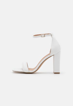 Białe sandały na wysokim obcasie z paskiem wokół kostki, z kwadratowym noskiem, gładką skórzaną powłoką, szerokim obcasem i wyściełaną wkładką dla wsparcia.