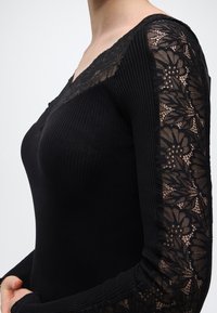 Μαύρο μακρυμάνικο τοπ με ραβδώσεις και φλοράλ δαντελένιες λεπτομέρειες στα μανίκια και το ντεκολτέ, φορεμένο από ένα άτομο που φαίνεται από τους ώμους μέχρι τους γοφούς.