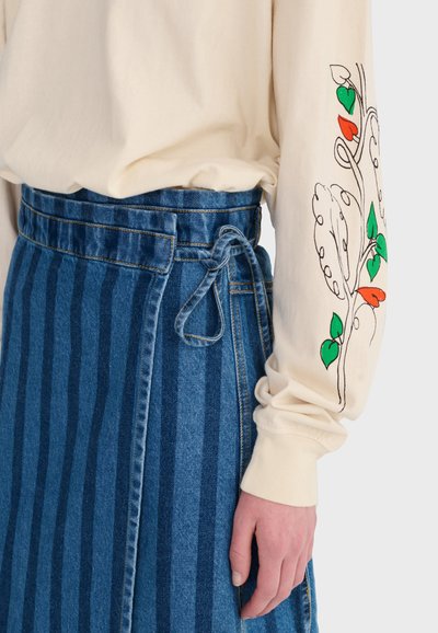 Person wearing a cream long-sleeve shirt with green and red leaf designs and a high-waisted blue striped denim skirt with a side tie.