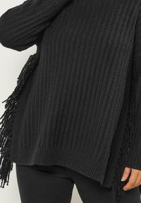 Czarny sweter z dzianiny o prążkowanej fakturze, z frędzlami na brzegach i luźnym kroju. W połączeniu z czarnymi spodniami.