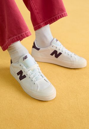 Białe buty sportowe New Balance z czarnym logo "N" noszone z białymi skarpetkami i czerwonymi spodniami na żółtej podłodze.