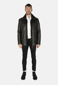 Svart läderjacka med hög krage, flera dragkedjefickor och strukturerad yta, kombinerad med svarta slim-fit byxor och svarta skor.