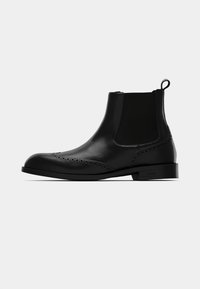 Cizmă Chelsea din piele neagră cu detalii brogue, panouri laterale elastice și o buclă de prindere pe o talpă plată, pe un fundal simplu.