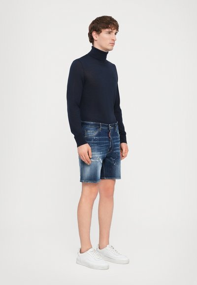 Marineblauer Rollkragenpullover, Jeansshorts mit ausgefransten Kanten und leichter Abnutzung, kombiniert mit weißen Turnschuhen, vor einem schlichten Hintergrund stehend.
