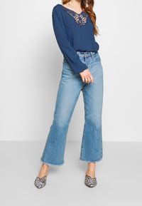 Marinebluse mit Spitzenbesatz am Ausschnitt, kombiniert mit hellblauen Schlagjeans mit rohem Saum und gemusterten Slippern mit schwarz-weißen Streifen.