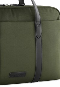 Olivgrüne Tasche mit strukturiertem Stoffoberfläche, schwarzen Lederakzenten und einem Logo-Patch, Reißverschlussverschluss und kurzen Griffen.