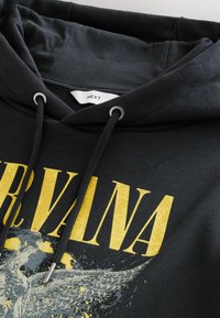 Melnā kapuce ar lielu dzeltenu tekstu "NIRVANA" un grafisku attēlu ar pelēkiem akcentiem. Iekļautas melnas saites un sudraba detaļas.