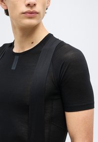 Νέος άνθρωπος που φοράει μαύρο υφασμάτινο κοντομάνικο πουκάμισο με φαρδείς τιράντες στους ώμους, στραμμένος ελαφρώς προς τα αριστερά, σε ανοιχτό φόντο.