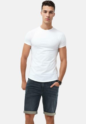 Weißes Kurzarm-T-Shirt kombiniert mit dunklen Jeansshorts mit umgeschlagenen Säumen. Das Model steht mit einer Hand in der Tasche vor einem neutralen Hintergrund.
