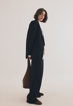 Schwarzer, übergroßer Blazer mit weit geschnittenen Hosen, darunter sichtbar ein beiger Pullover sowie eine braune Tote-Tasche. Das Modell steht vor einem neutralen Hintergrund.