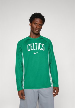 Mladý muž v zelenej dlhorukávovej športovej košeli Celtics a sivých šortkách stojí na jednoduchom svetlom pozadí.