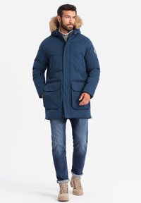 Blå isolerad parkas med pälskantad huva, stora framfickor, knäppning med knappar, bärs över en grå tröja och blå jeans.