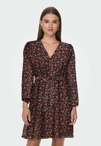 Donna che indossa un abito floreale nero con scollo a V, maniche lunghe e una cintura legata in vita, in piedi davanti a uno sfondo chiaro e uniforme.