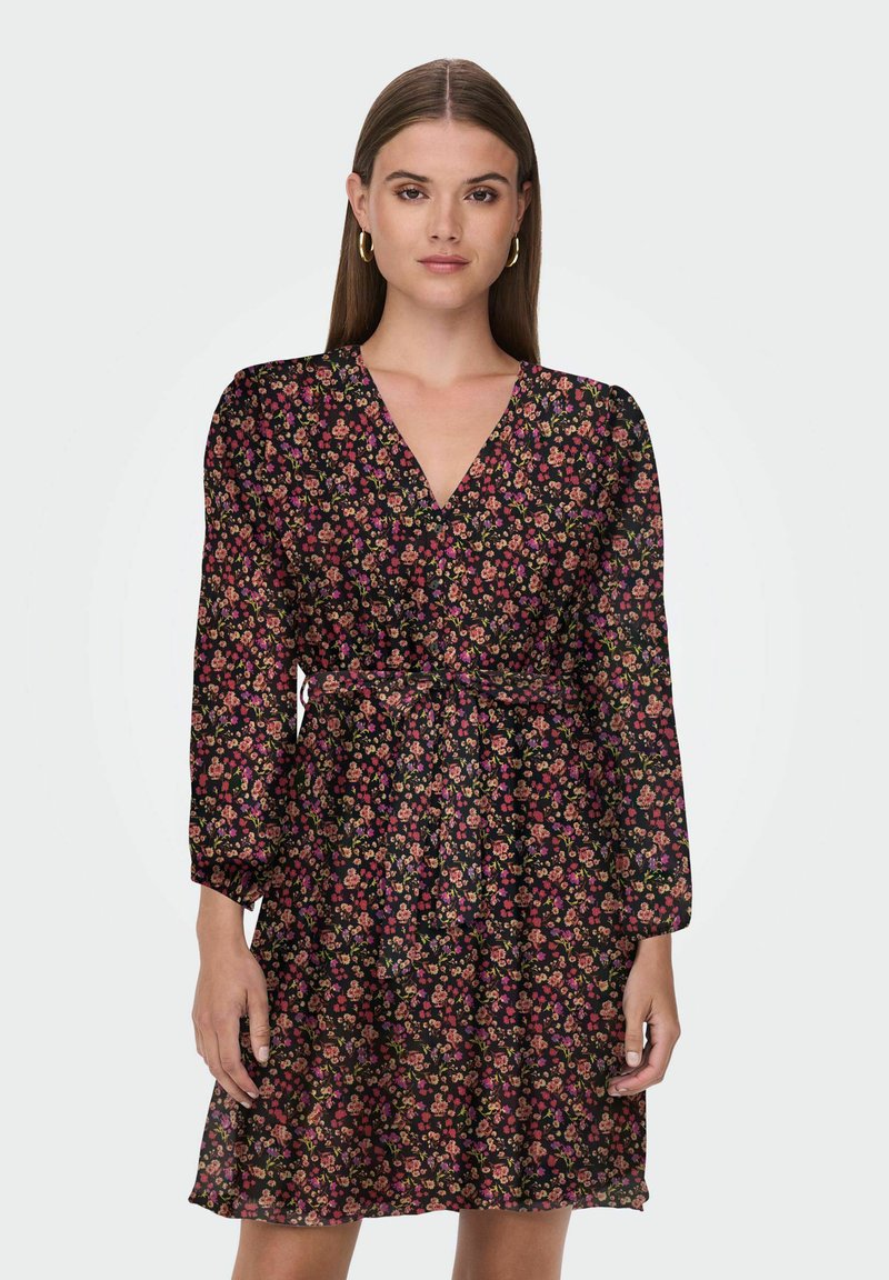 Donna che indossa un abito floreale nero con scollo a V, maniche lunghe e una cintura legata in vita, in piedi davanti a uno sfondo chiaro e uniforme.