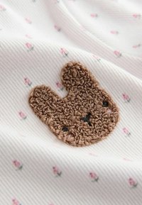 Light fabric with a white base featuring small pink flower patterns, accented by a brown crocheted bunny face with black eyes.