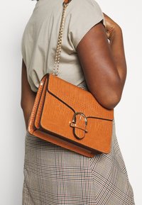 Beżowa torebka ze skóry tłoczonej w wzór krokodyla, z złotym łańcuszkowym paskiem, o usztywnionym kształcie i detalem z klamrą. Dopasowana do spódnicy w kratę.