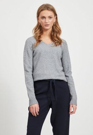 Pulover gri cu guler în V, realizat dintr-un material tricotat moale, cu manșete ribbate și o croială lejeră. Asortat cu pantaloni închiși la culoare, cu șnur.