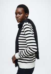 Maglione a righe bianche e nere con maniche lunghe, abbinato a una sciarpa nera unita avvolta intorno al collo. Capelli corti e testurizzati visibili.