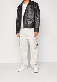 Černá kožená motorkářská bunda se zipy, pod ní bílá tričko, světlé béžové cargo kalhoty s bočními kapsami a bílé tenisky.