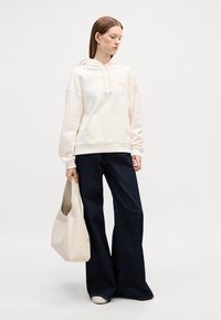 Felpa crema con tasca frontale e lacci, abbinata a jeans ampi scuri. Il modello tiene una borsa hobo beige chiaro. Design e trame minimal.