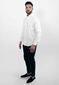 Witte lange mouwen overhemd met knopen, donkere slim-fit broek, zwarte instap schoenen. Model staat tegen een plain witte achtergrond.