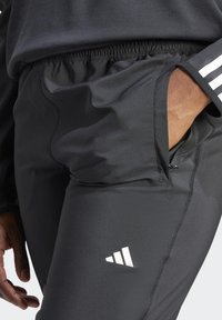 Czarne spodnie sportowe z elastycznym pasem, boczną kieszenią i białym logo Adidasa. Gładka faktura o delikatnym połysku i kontrastowym przeszyciu.