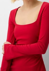 Rød tætsiddende top med lange ærmer, firkantet udskæring og klokkeformede manchetter. Fremstillet af glat, strækbart stof med en elegant overflade.