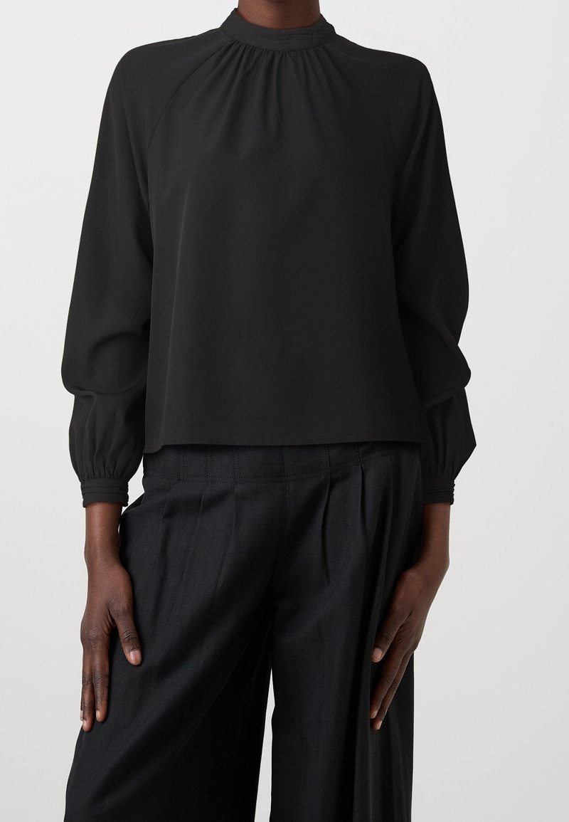Femme portant un chemisier noir à manches longues avec un col froncé et un pantalon noir plissé et ample sur un fond uni.