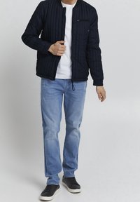 Giacca bomber trapuntata navy con tasca anteriore con zip, colletto e polsini a coste, abbinata a jeans azzurri chiari e sneakers nere.