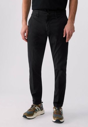 Homme portant un pantalon noir slim et des baskets multicolores, debout devant un fond clair uni.