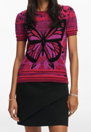 Mujer que lleva un suéter de manga corta rosa y rojo con un gran diseño de mariposa negro y rosa, combinado con una falda negra asimétrica.
