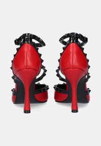 Zapatos de tacón alto de cuero rojo con correas negras con tachuelas y tacones puntiagudos, que presentan un diseño elegante y una textura suave.