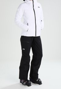 Weiß isolierte Jacke mit Kapuze und schwarzem Reißverschluss, kombiniert mit schwarzen Skihosen. Auffällige Details sind Taschen und Logos. Skischuhe sind inklusive.