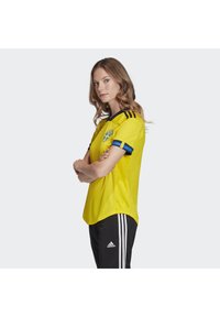 Gul atletiktrøje med blå accenter og svensk emblem. Korte ærmer, rund krave og meshstruktur. Parret med sorte bukser med tre striber.