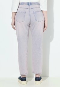 Persona che indossa jeans viola chiaro e sneakers blu scuro, in piedi di fronte a un muro bianco con le mani rilassate ai lati.